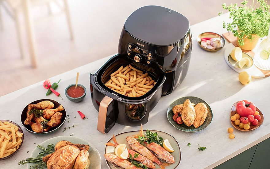An air fryer filled with fries sits on a table surrounded by plates of cooked food and fresh ingredients.