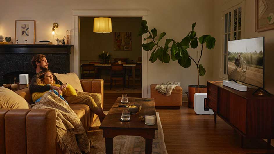 Een woonkamer met een bruine leren bank, houten tafel en tv. Er staat een grote plant en een open doorgang naar een eetkamer.