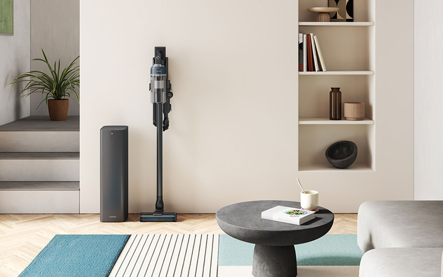 Intérieur clair avec aspirateur noir, support et table ronde grise. Étagère murale avec livres et décorations. Tapis rayé bleu et blanc.