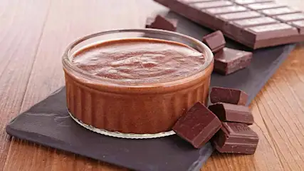 Een glazen schaaltje met chocolademousse staat op een zwart bord, met stukken chocolade en een reep op een houten tafel.