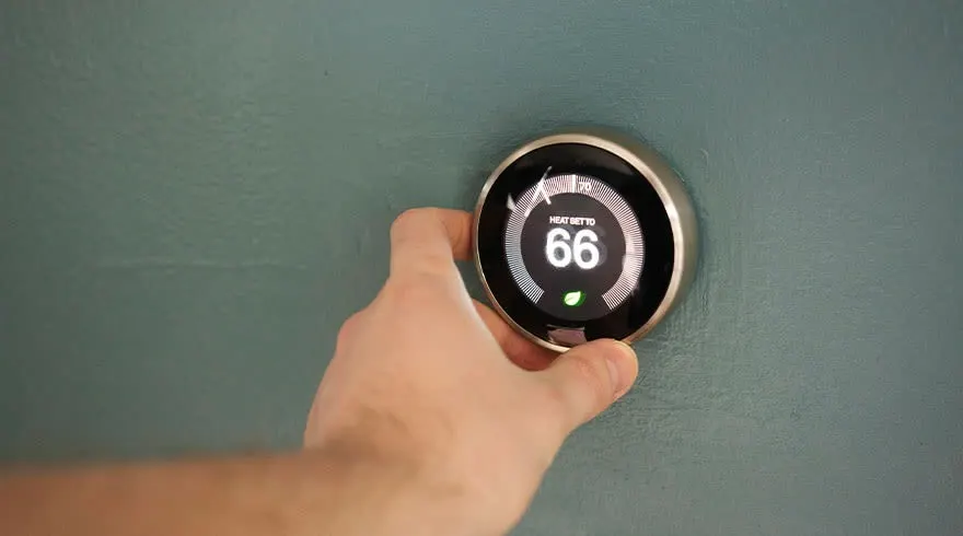 Een hand past een ronde, digitale thermostaat aan op een blauwgroene muur. De thermostaat toont 66 graden.