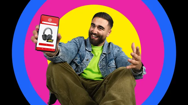 Zittende persoon in denimjas toont glimlachend een smartphone met een aanbieding voor koptelefoons.