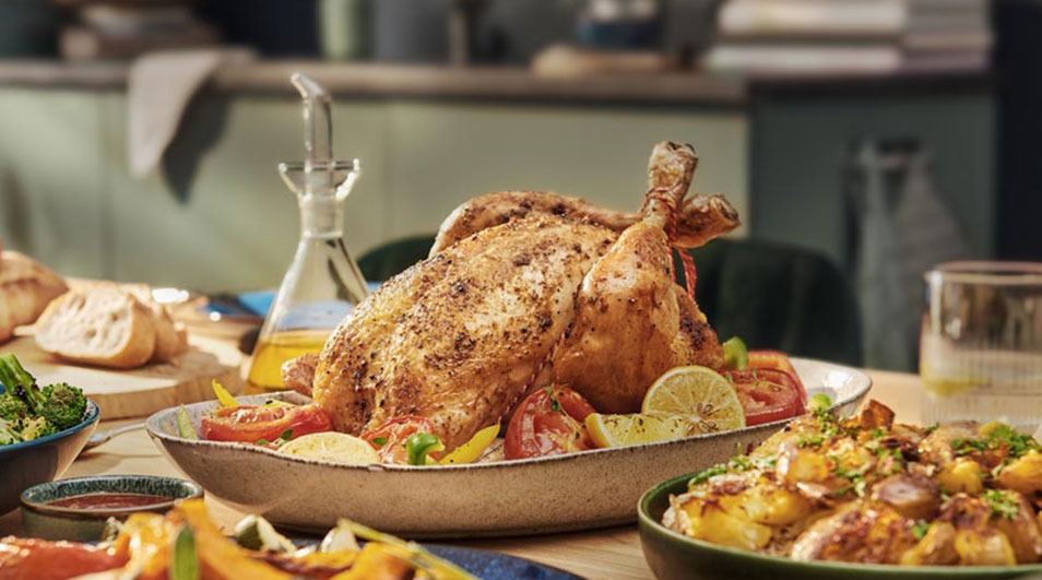 A table displays a roast chicken on a platter with tomatoes and lemon, surrounded by bowls of potatoes, vegetables, bread, and a bottle of oil.