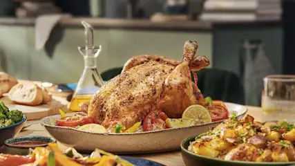 Un poulet rôti doré sur un plat avec des tomates et du citron, sur une table avec des accompagnements variés. Carafe d'huile en arrière-plan.