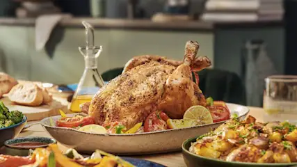 Gegrilde kip op een schaal met citroen en tomaat, met brood en aardappelen, op een houten tafel in een keuken.
