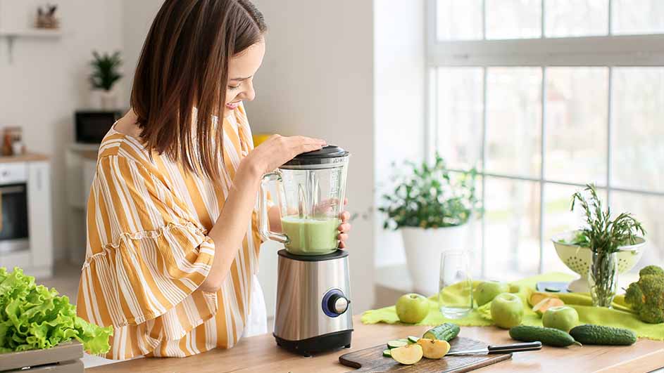Une cuisine claire avec un mixeur rempli d'un liquide vert, entouré de fruits et légumes frais sur une table en bois.