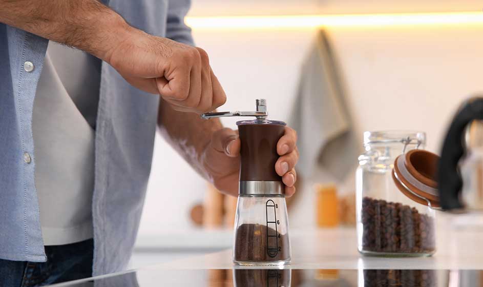 Een persoon maalt koffiebonen met een handmatige molen boven een glazen pot. Op het aanrecht staat een voorraadpot met bonen en een borstel.