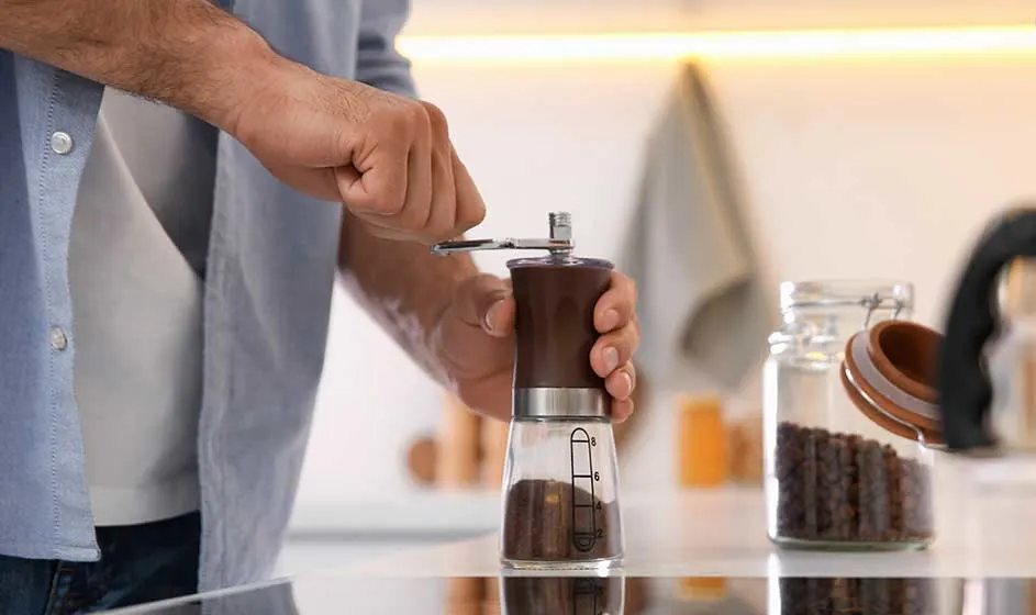 Een persoon maalt koffiebonen met een handmatige molen boven een glazen pot. Op het aanrecht staat een voorraadpot met bonen en een borstel.
