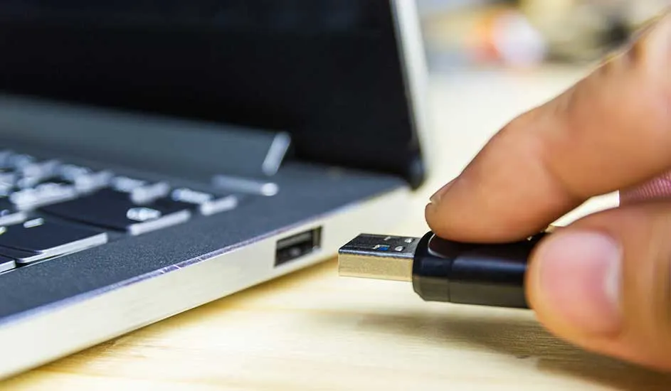 Een hand steekt langzaam een zwarte USB-stick in de zijkant van een open laptop op een houten bureau.