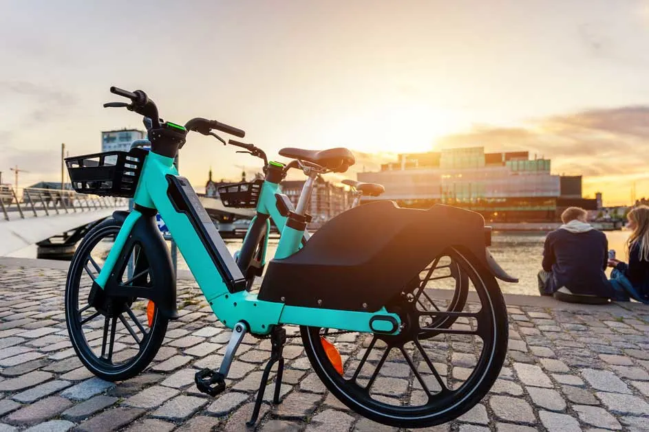 Twee turquoise elektrische fietsen van Tier staan op een kade bij zonsondergang, met mandje voorop en moderne gebouwen op de achtergrond.