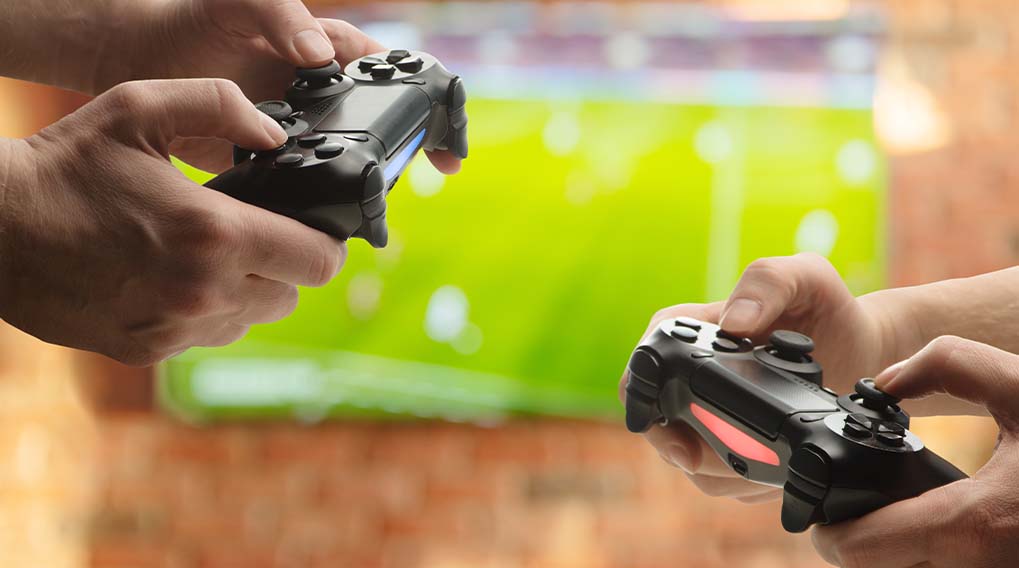 Deux mains tiennent des manettes noires. En arrière-plan, un écran vert affiche un jeu.