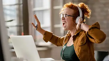 Een witte laptop staat open voor een raam. Een bruin hemd en witte hoofdtelefoon zijn zichtbaar.