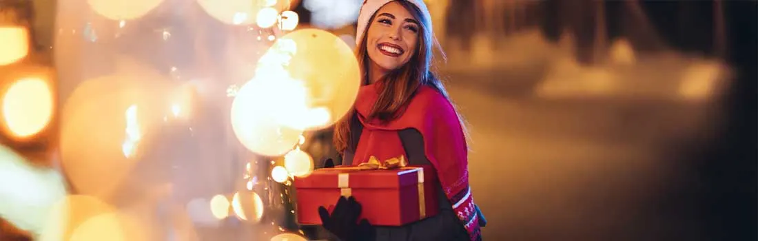 Een vrouw met een kerstmuts loopt vrolijk langs feestelijke lichtjes buiten en houdt een rood, goud gestrikt cadeautje vast.