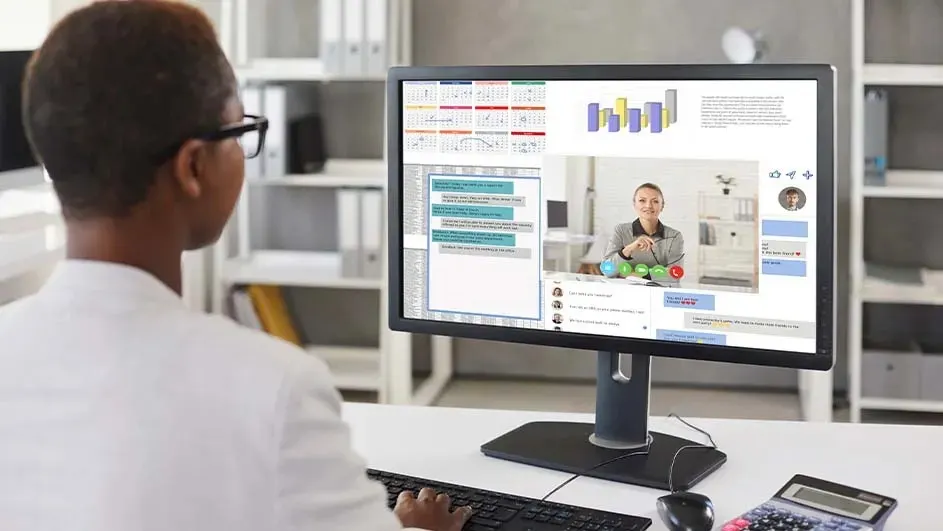 Iemand gebruikt een computer met grafieken en een video gesprek op het scherm, plus een rekenmachine op een wit bureau.