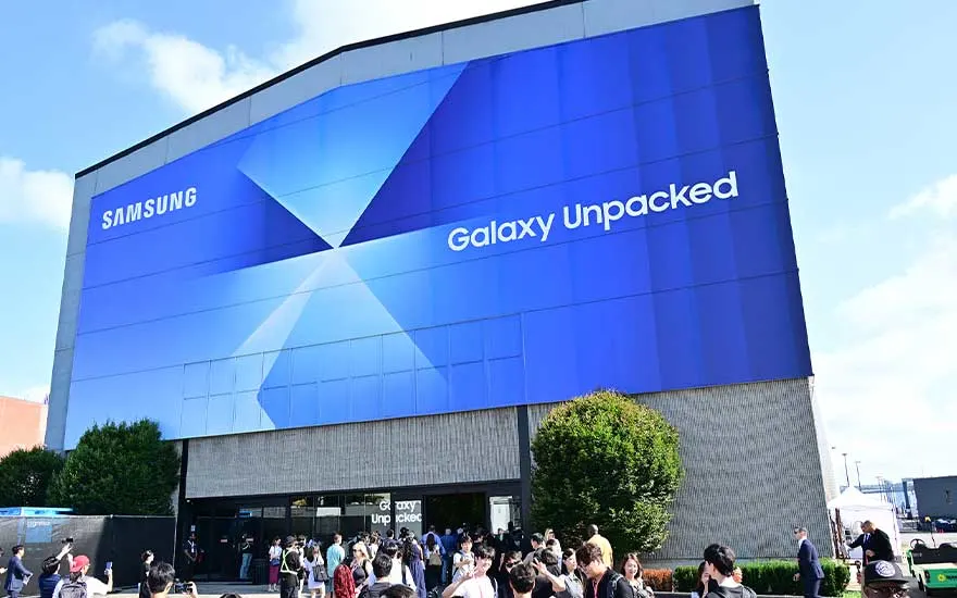 Een groot gebouw is bekleed met een blauwe Samsung Galaxy Unpacked-banner, terwijl bezoekers buiten bij de ingang samenkomen.