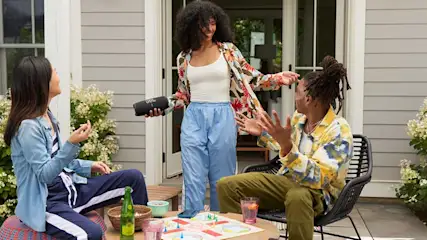Trois personnes à l'extérieur avec des boissons et un jeu de société sur une table, avec un haut-parleur noir.