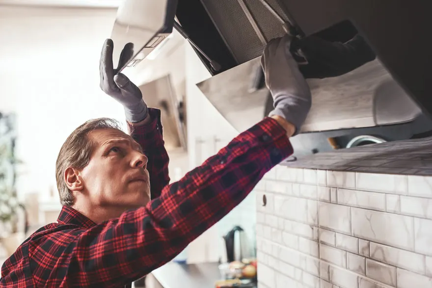 Un homme en chemise à carreaux installe une hotte de cuisine grise sur un mur carrelé blanc.