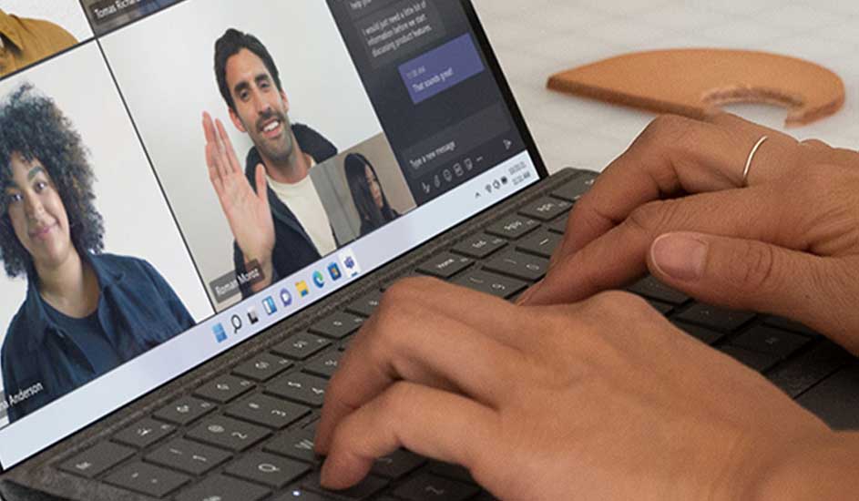 Op een laptop toont een scherm een video meeting met vier deelnemers. Iemand typt op het donkergrijze toetsenbord. Een koekje ligt op de achtergrond.