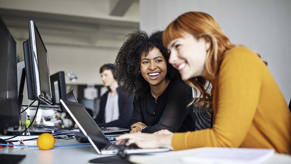 Kantoorbeeld met computers, een laptop en een sinaasappel op een wit bureau. Andere bureaus zichtbaar op de achtergrond.