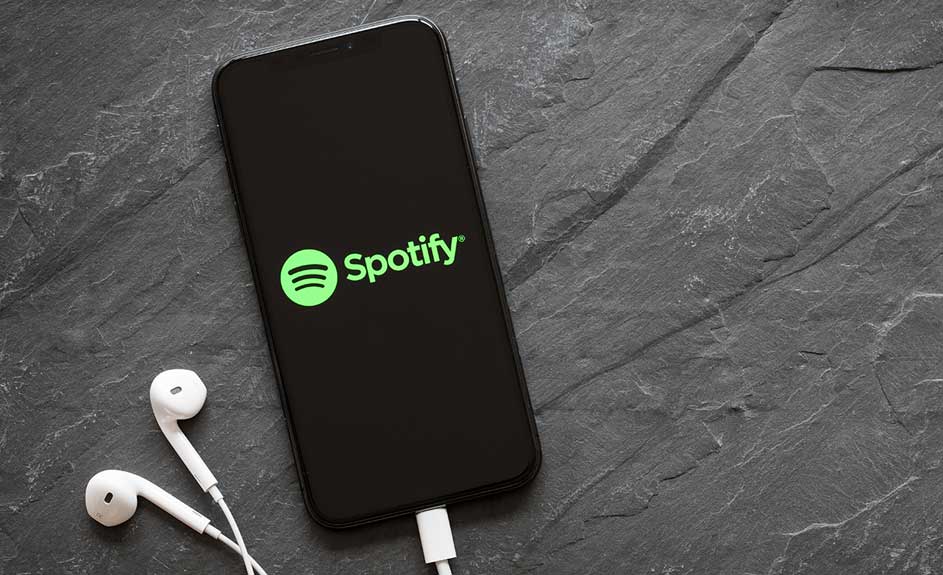 Un téléphone noir affiche Spotify avec des écouteurs blancs sur fond d'ardoise grise texturée.