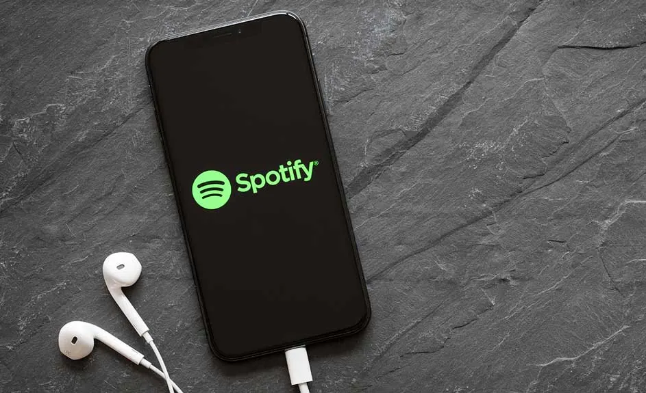 Un téléphone noir affiche Spotify avec des écouteurs blancs sur fond d'ardoise grise texturée.