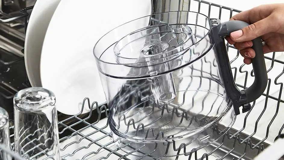 Un panier de lave-vaisselle en métal contient une assiette blanche, des verres et un récipient en plastique transparent avec une poignée noire.