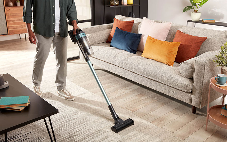 Un aspirateur bleu est utilisé sur un tapis beige dans un salon avec un canapé, des coussins colorés et une table basse noire.