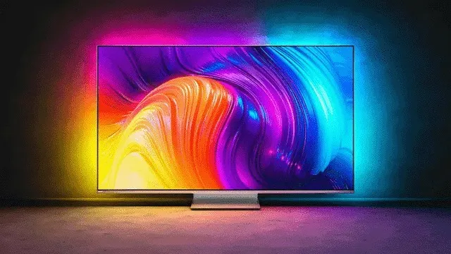 Een grote tv staat op een zilveren voet. Het scherm toont abstracte, golvende vormen in geel, oranje, paars en blauw. Het licht reflecteert op de muur.