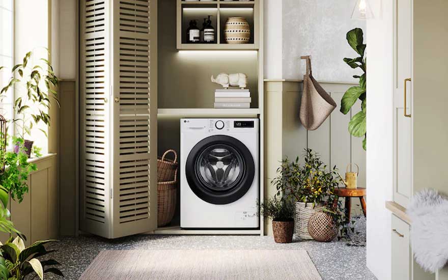 Compacte wasmachine in een lichte, decoratieve nis met houten luiken, manden, planten, planken en een rustige huiselijke sfeer.