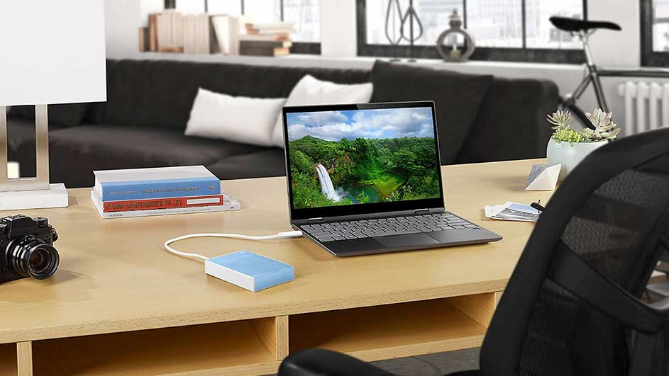 Bureau avec ordinateur portable affichant une cascade, un appareil photo, des livres et un disque dur externe. Canapé en arrière-plan.