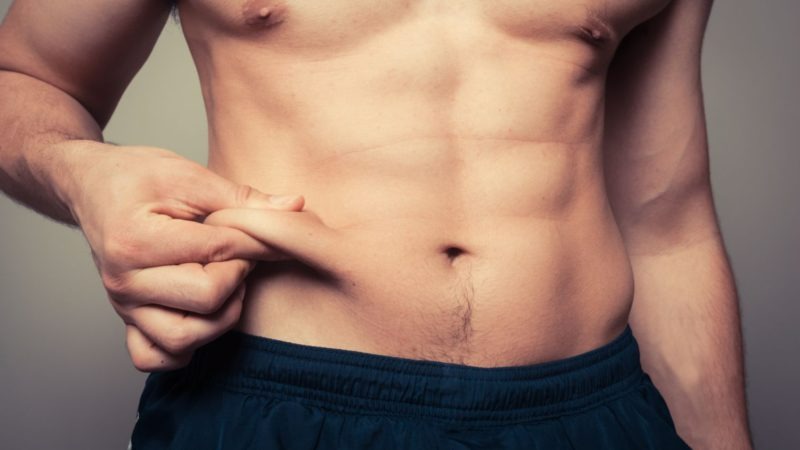 Un torse nu avec une main pinçant la peau du ventre, sur un fond gris clair. Pantalon bleu visible.