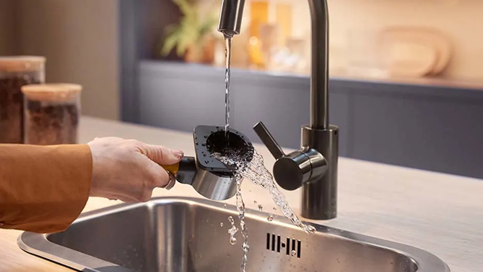 L'eau coule d'un robinet noir dans un évier en acier. Une main tient un filtre à café sous l'eau. Bocaux en verre visibles en arrière-plan.