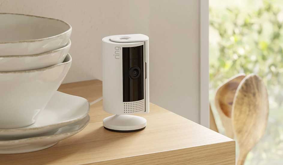 Une caméra de surveillance Ring blanche est posée sur une table en bois à côté de bols. Son design est moderne et compact.