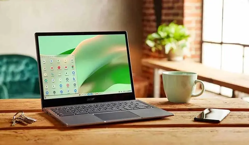 Un ordinateur portable gris est sur une table en bois avec des clés, un téléphone et une tasse verte. En arrière-plan, une plante et une fenêtre.