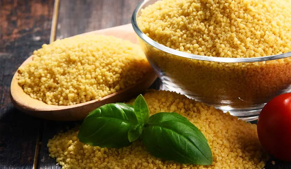 Couscous jaune dans un bol en verre, une cuillère en bois, sur une table en bois avec du basilic et une tomate rouge.