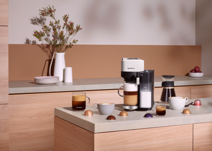 Koffiezetapparaat op aanrecht met kopjes en capsules, plus bloemenvaas. Achtergrond is beige en wit.