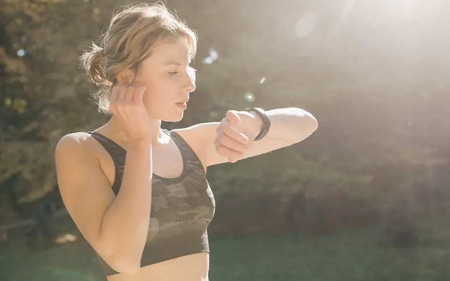 Une personne porte un soutien-gorge de sport et une montre intelligente à l'extérieur, avec un arrière-plan de verdure et de soleil.