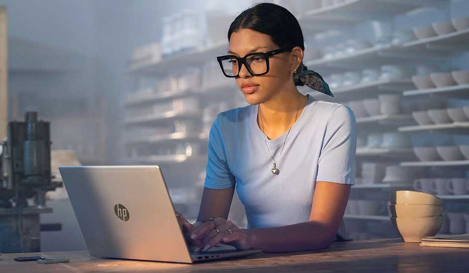 Femme portant des lunettes travaille sur un ordinateur HP à une table en bois, entourée d’étagères remplies de céramiques.