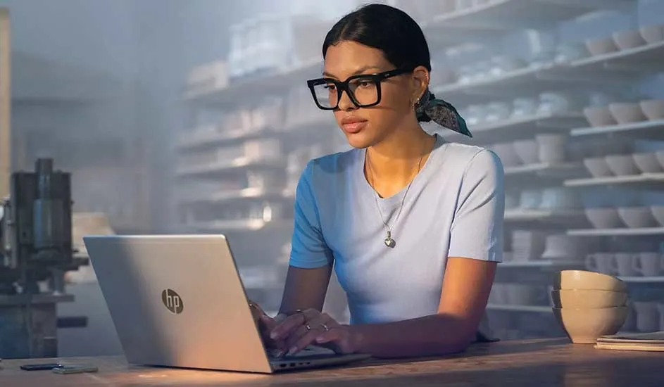 Vrouw met bril werkt gefocust op HP‑laptop aan houten tafel, omringd door schappen vol keramiek in zacht verlichte werkplaats.