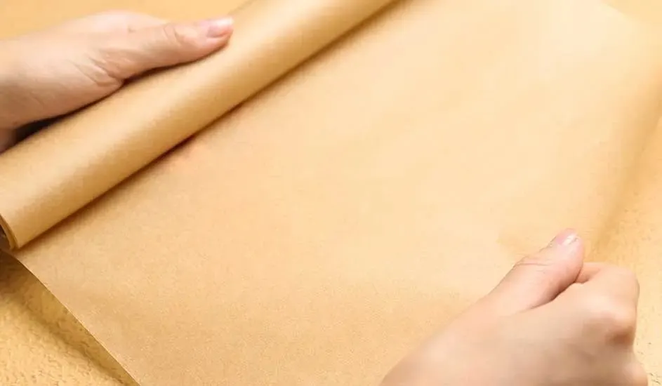 Twee handen rollen een vel bakpapier uit op een tafel, merk is niet zichtbaar.