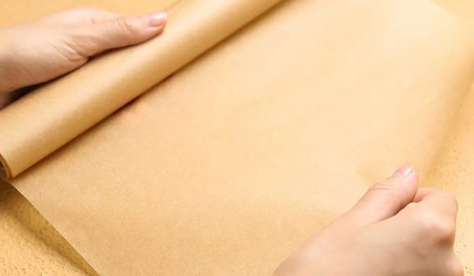 Hands unroll a sheet of baking parchment on a flat surface, ready for use in cooking or baking in the kitchen.