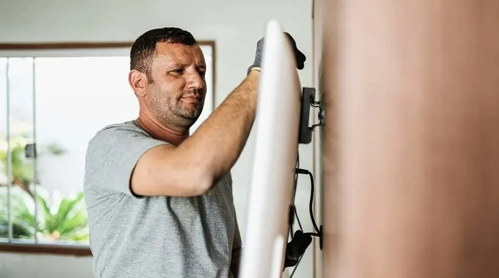 Een man in een grijze T-shirt monteert een witte televisie aan een beige muur met een raam op de achtergrond.