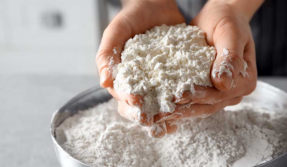 Twee handen houden een hoop wit meel boven een metalen kom vol met meer meel.