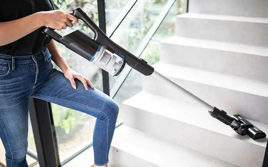 Een zwarte stofzuiger wordt gebruikt op een witte trap naast een raam, met een tweede opzetstuk op een trede.