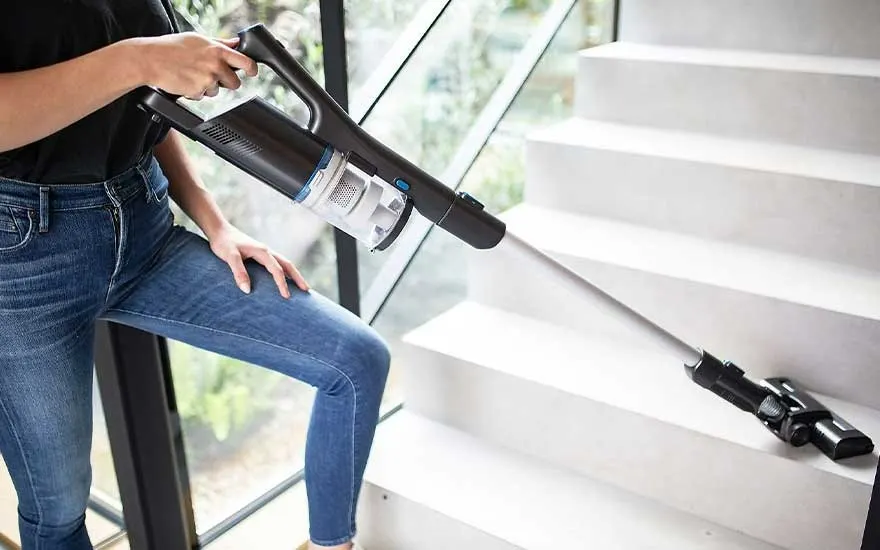 Une personne tient un aspirateur noir et blanc près d'un escalier blanc, avec un embout sur une marche.
