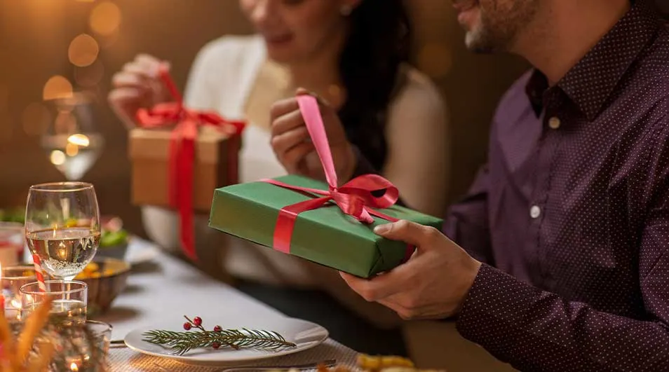 Un couple assis à une table joliment décorée échange des cadeaux soigneusement emballés, dans une ambiance chaleureuse et festive.