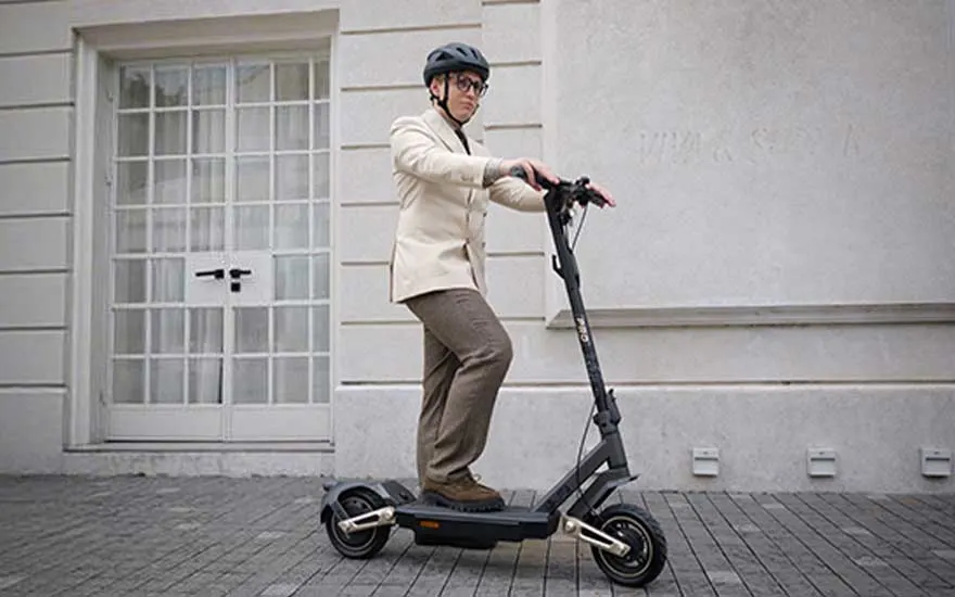 Een jongere rijdt op een kleurrijke elektrische step door een plas water voor een moderne woning, met warm avondlicht.