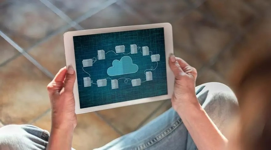 Tablet displays a cloud and file icons on a blue screen. Hands hold the tablet. Background shows a tiled floor.