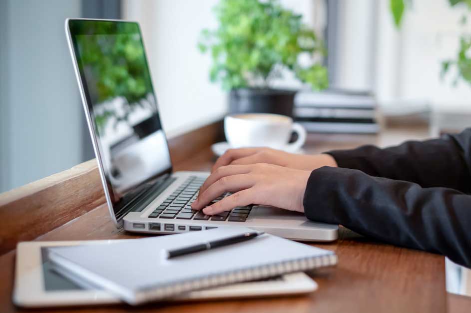 Een zilveren laptop op een houten tafel met een notitieboek, een pen en een kop koffie op de achtergrond.