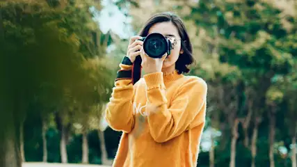 Une personne prend une photo avec un appareil photo noir. L'arrière-plan montre des arbres verts.